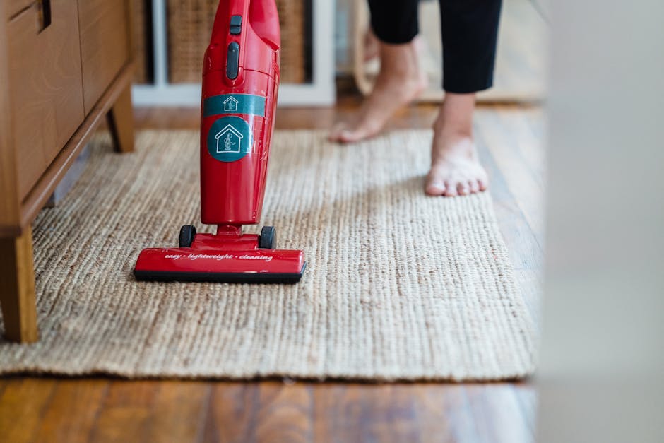 Close-up of a person cleaning a beige textured carpet with a red handheld vacuum cleaner, positioned on a wooden floor near a wooden piece of furniture. The individual's feet are visible, clad in blac