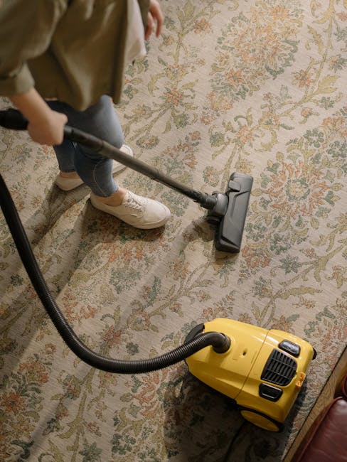 Close-up of a person cleaning a beige textured carpet with a red handheld vacuum cleaner, positioned on a wooden floor near a wooden piece of furniture. The individual's feet are visible, clad in blac