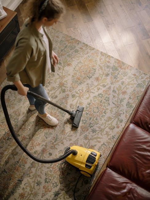 A young boy vacuuming a beige sofa in a bright living room with white walls and large windows, while an older man with grey hair and a beard, dressed in a light grey t-shirt and blue jeans, sits on th